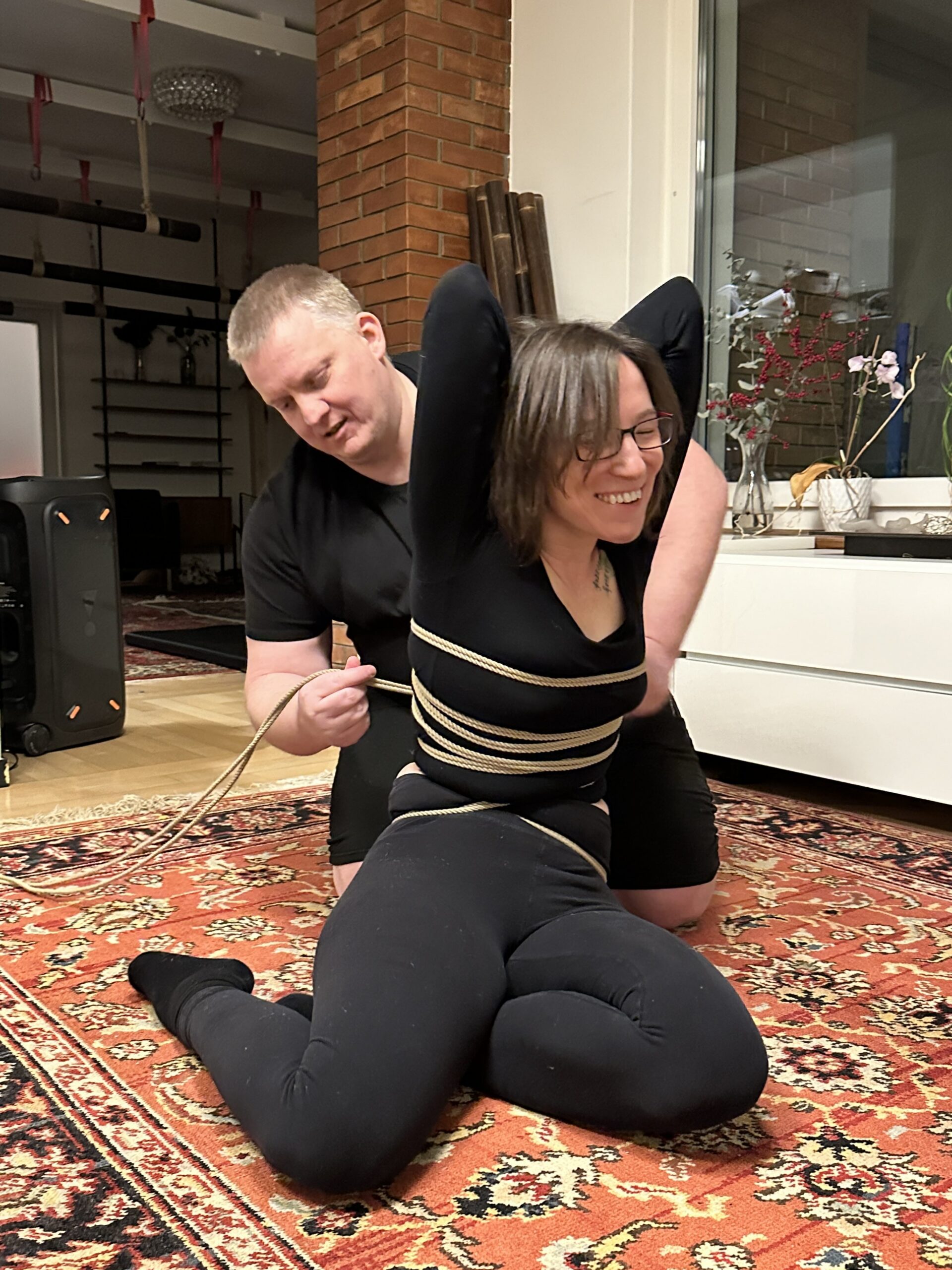 Christian kniet hinter Jay auf dem Boden und fesselt sie mit Jute Seilen in ersten Shibari Schritten. Jay lächelt.