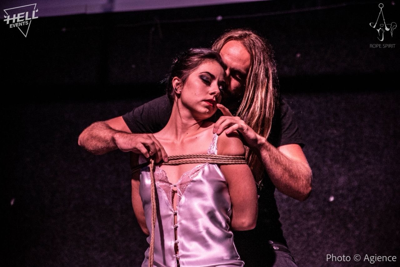 Shibari Rope Performance Aschaffenburg 3 Ein Rigger zieht Seil über die Brust einer Frau während einer Shibari Performance.